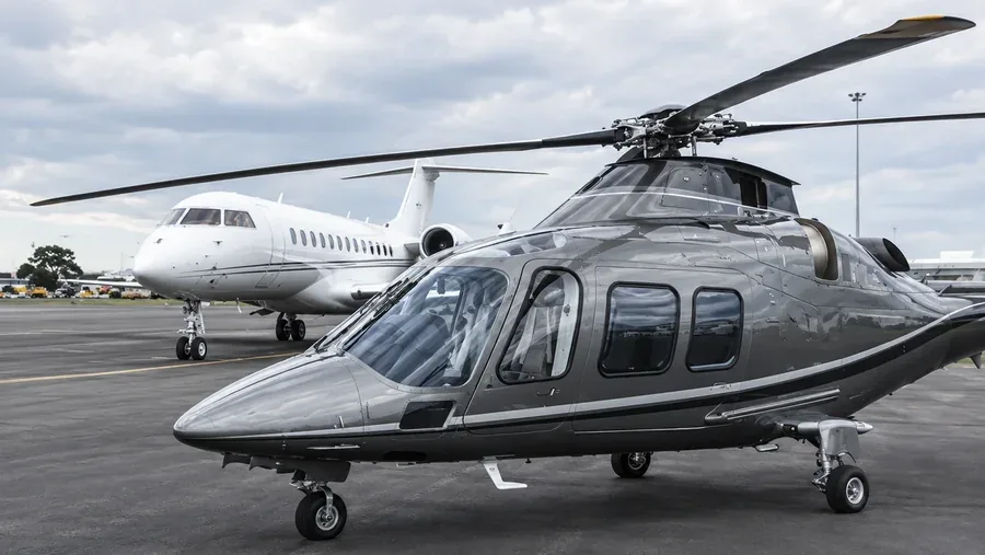 A grey Agusta 109 helicopter on the apron with a business jet behind it