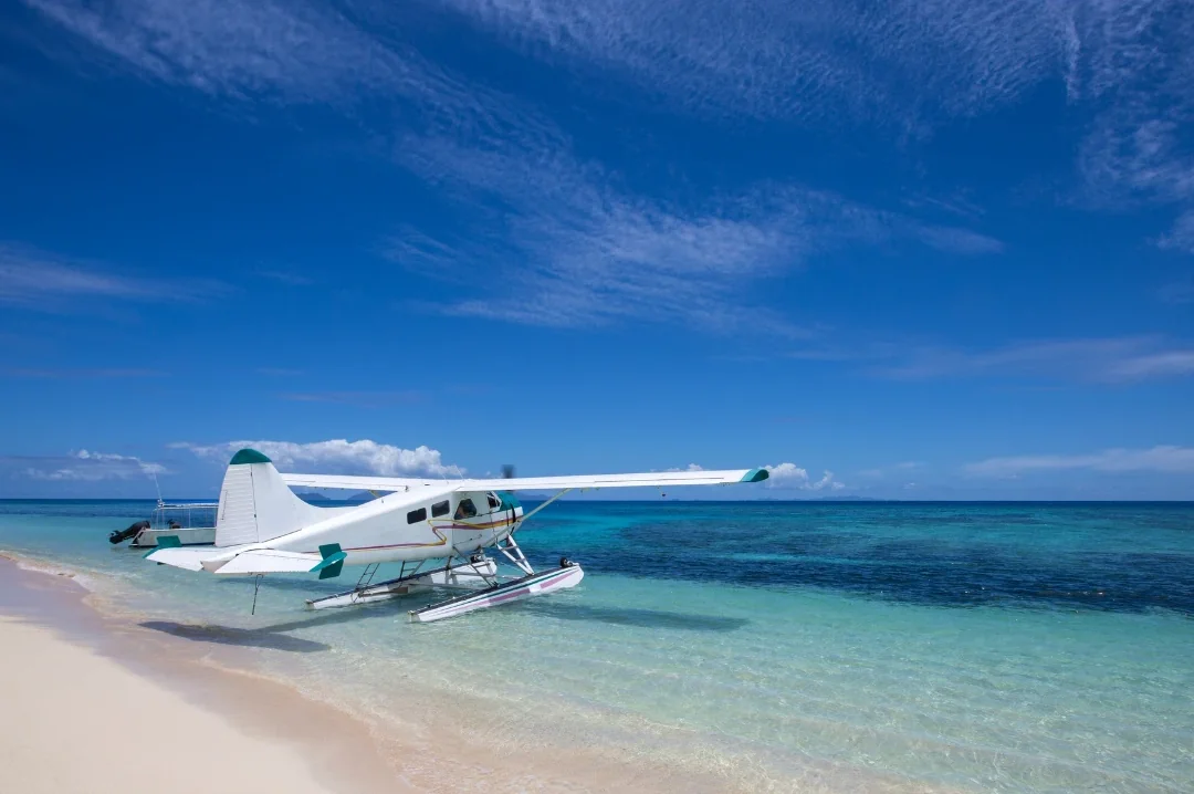 A white seaplane on floats at the edge of a white sand beach with turquoise water behind it